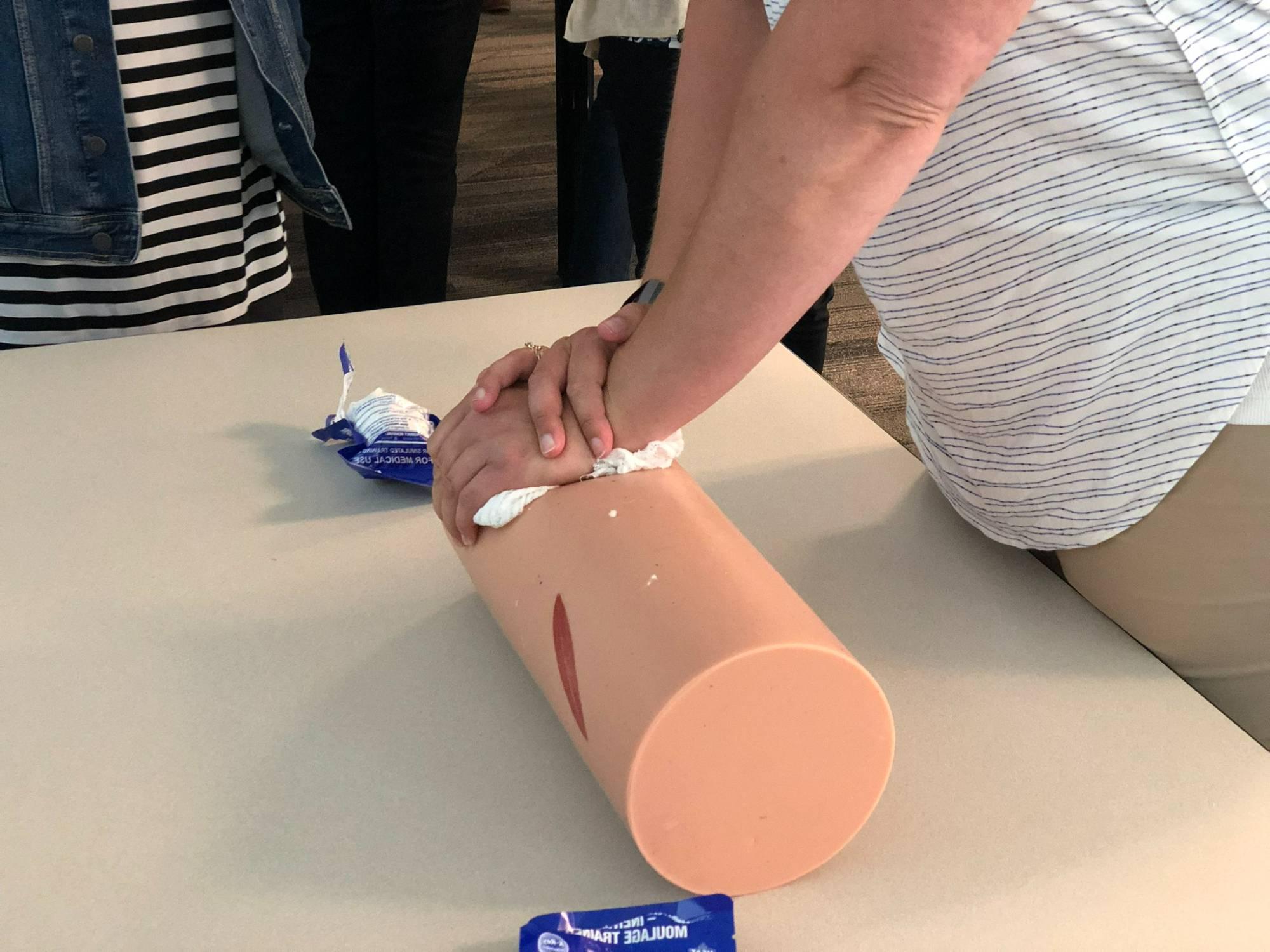 White woman's arms and hands crossed over a cylindrical item meant to simulate a person's leg, packing a wound with gauze